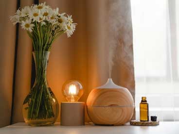 A glass vase of white flowers beside a lit bulb and a wooden essential oil diffuser releasing mist on a table.