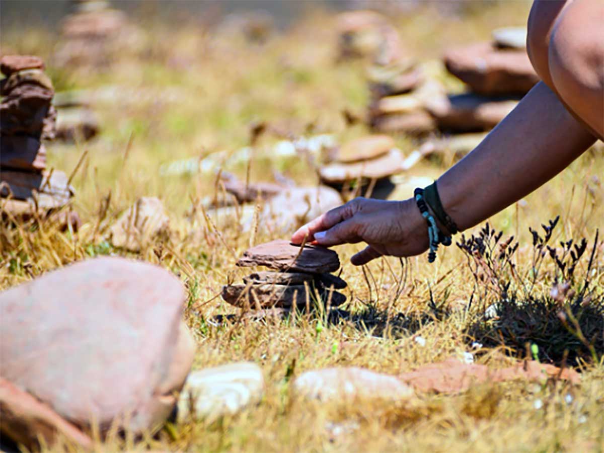 A person stacking flat stones outdoors in a grassy area with small rock piles in the background