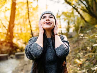 A smiling woman standing in a sunlit forest with her hands near her ears