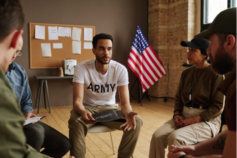 A group therapy or discussion session with a man wearing an "Army" t-shirt speaking to others seated in a circle, with an American flag in the background.