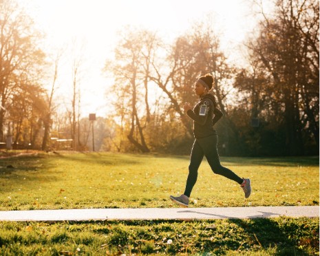 A woman jogging alone on a park path during golden hour, surrounded by trees with autumn foliage.